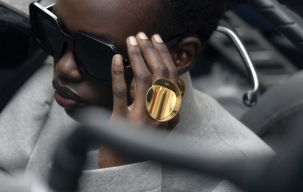 Close-up of a black adult woman wearing sunglasses and gold rings on her hand, with a grey sweater, and dark background.