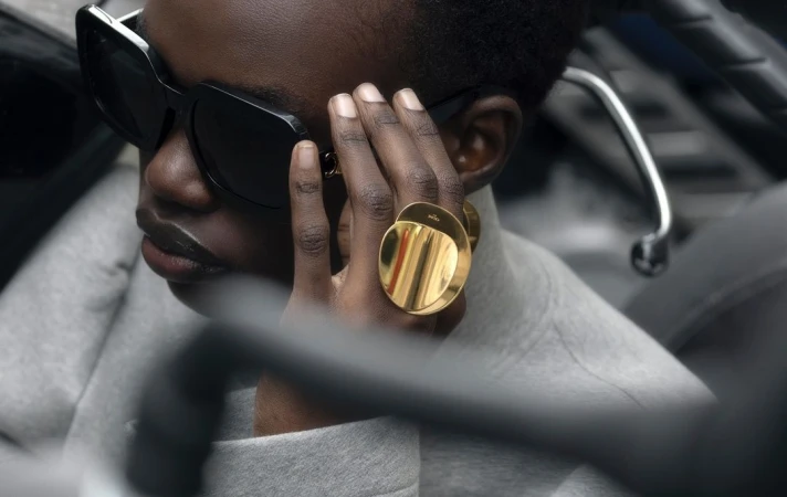 Close-up of a black adult woman wearing sunglasses and gold rings on her hand, with a grey sweater, and dark background.