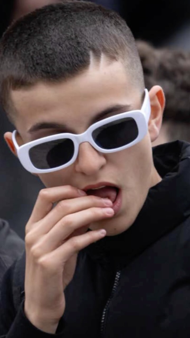 Portrait of a young adult with a buzz cut and white-frame glasses, biting a fingernail, wearing a black jacket, and standing outside.