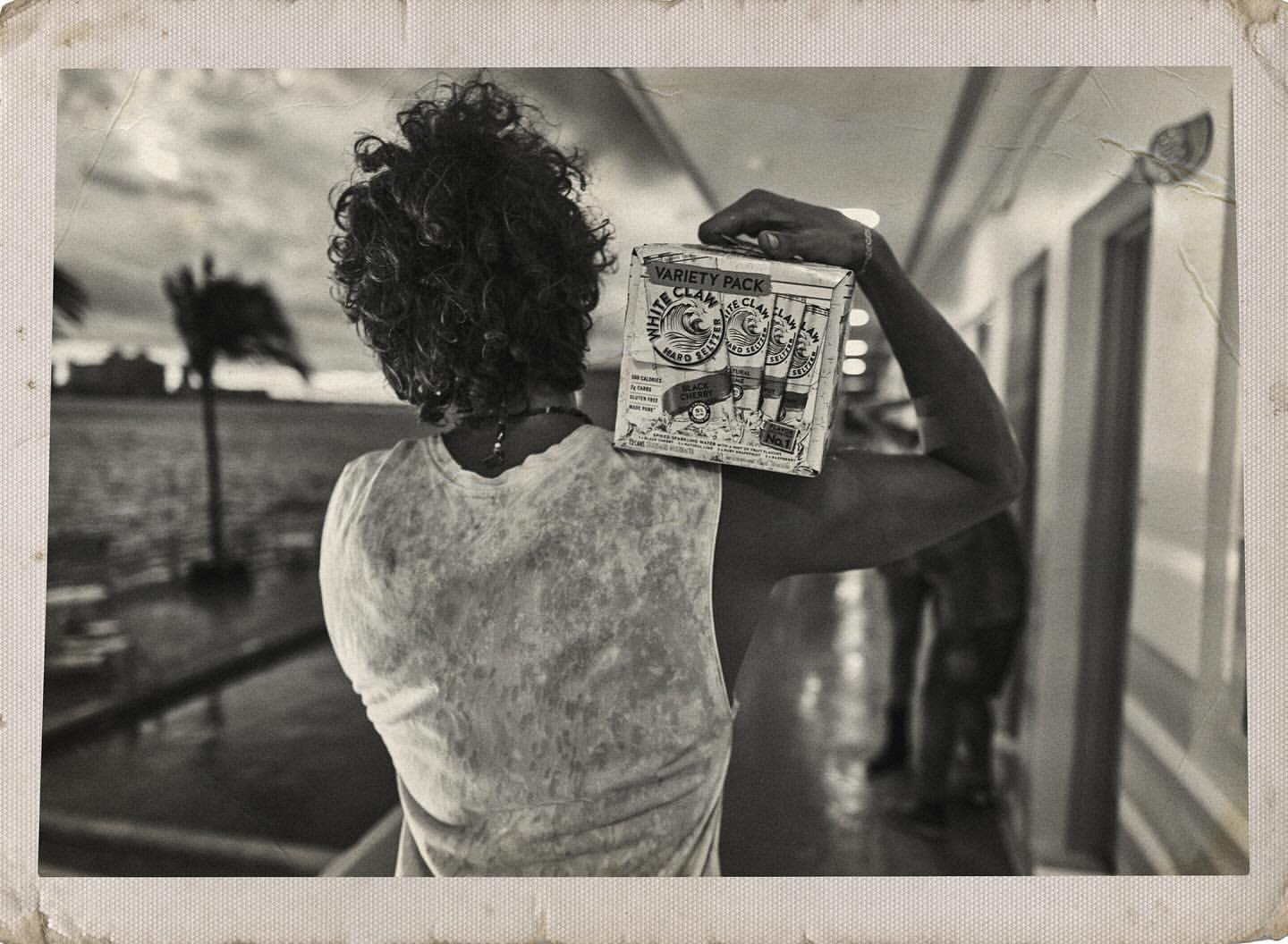 Monochrome photo of a person carrying a box of White Claw Variety Pack by the water with sparse trees, creating a vintage aesthetic.