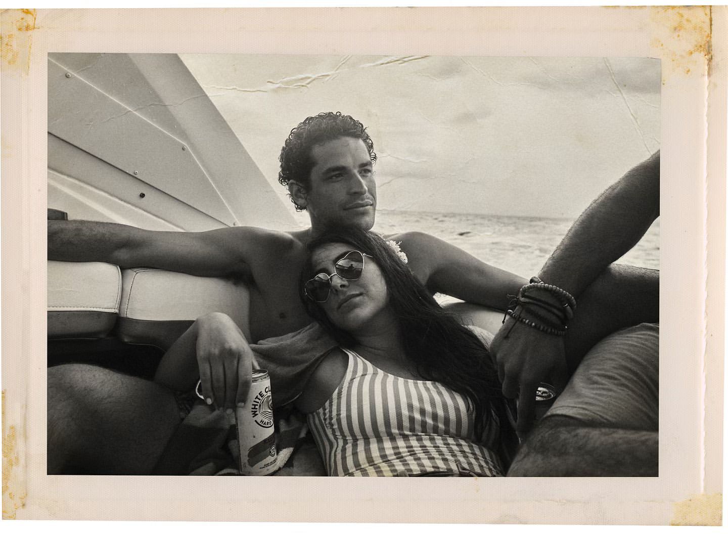 Monochrome photo of a man and woman relaxing on a boat in the ocean, the woman in sunglasses and a striped top, the man shirtless.