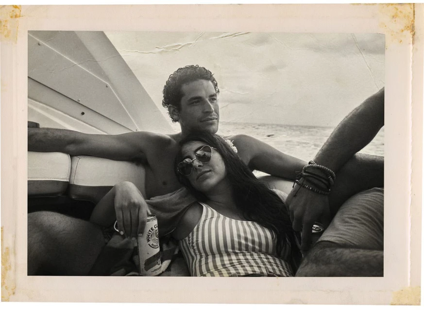 Monochrome photo of a man and woman relaxing on a boat in the ocean, the woman in sunglasses and a striped top, the man shirtless.