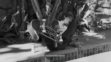 Black and white photo of a skater falling backwards while attempting a trick near a concrete pool with leafy plants in the background.