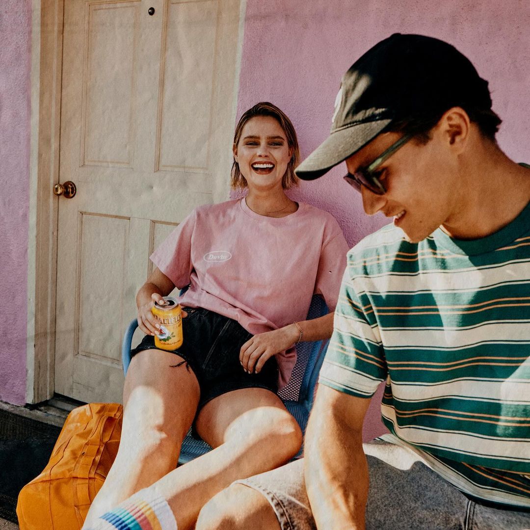 A smiling young woman in a pink t-shirt sits next to a young man wearing a striped shirt and baseball cap outdoors in soft lighting.