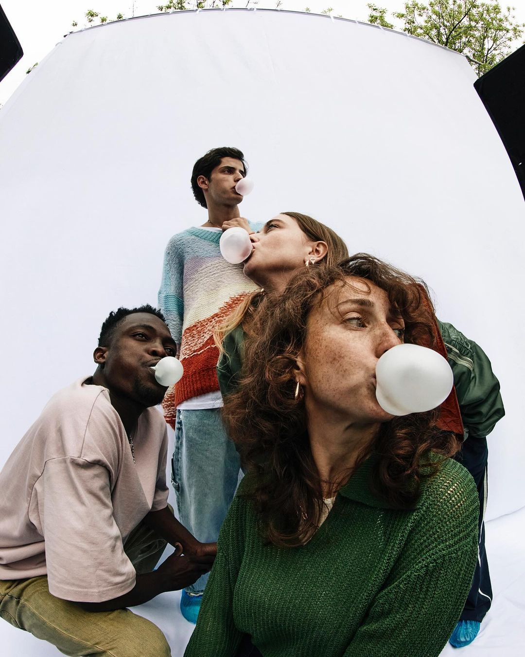 Four diverse people inflate pink bubblegum bubbles in a close-up studio shot with bright, even lighting and youthful energy.