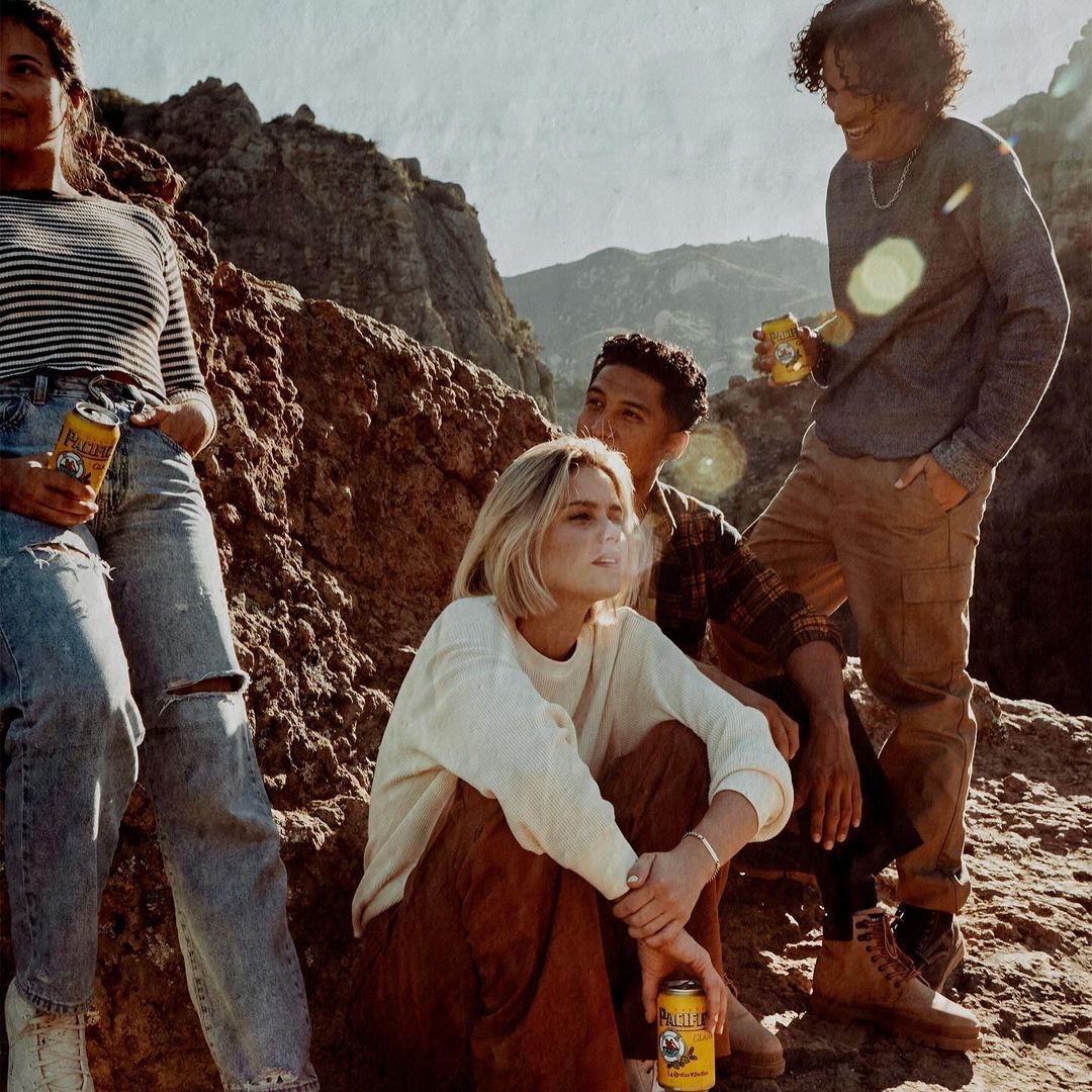 Group of four young adults pose with beer cans on a rocky hillside against a mountain backdrop on a sunny day. Lifestyle, outdoor.