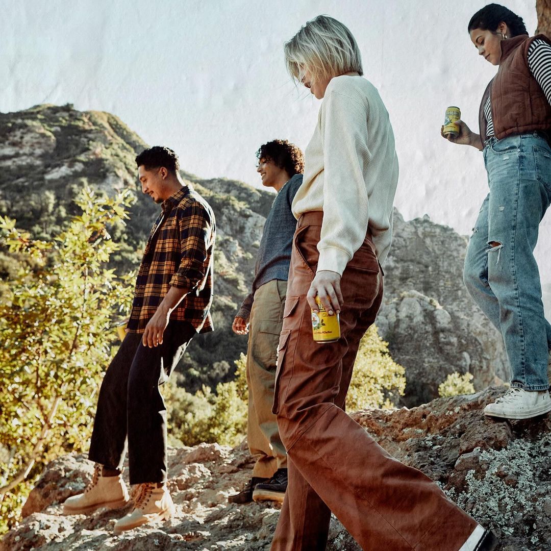 A group of four young adults hiking on a mountain trail in casual clothing, bathed in natural sunlight, with a mountain range backdrop.
