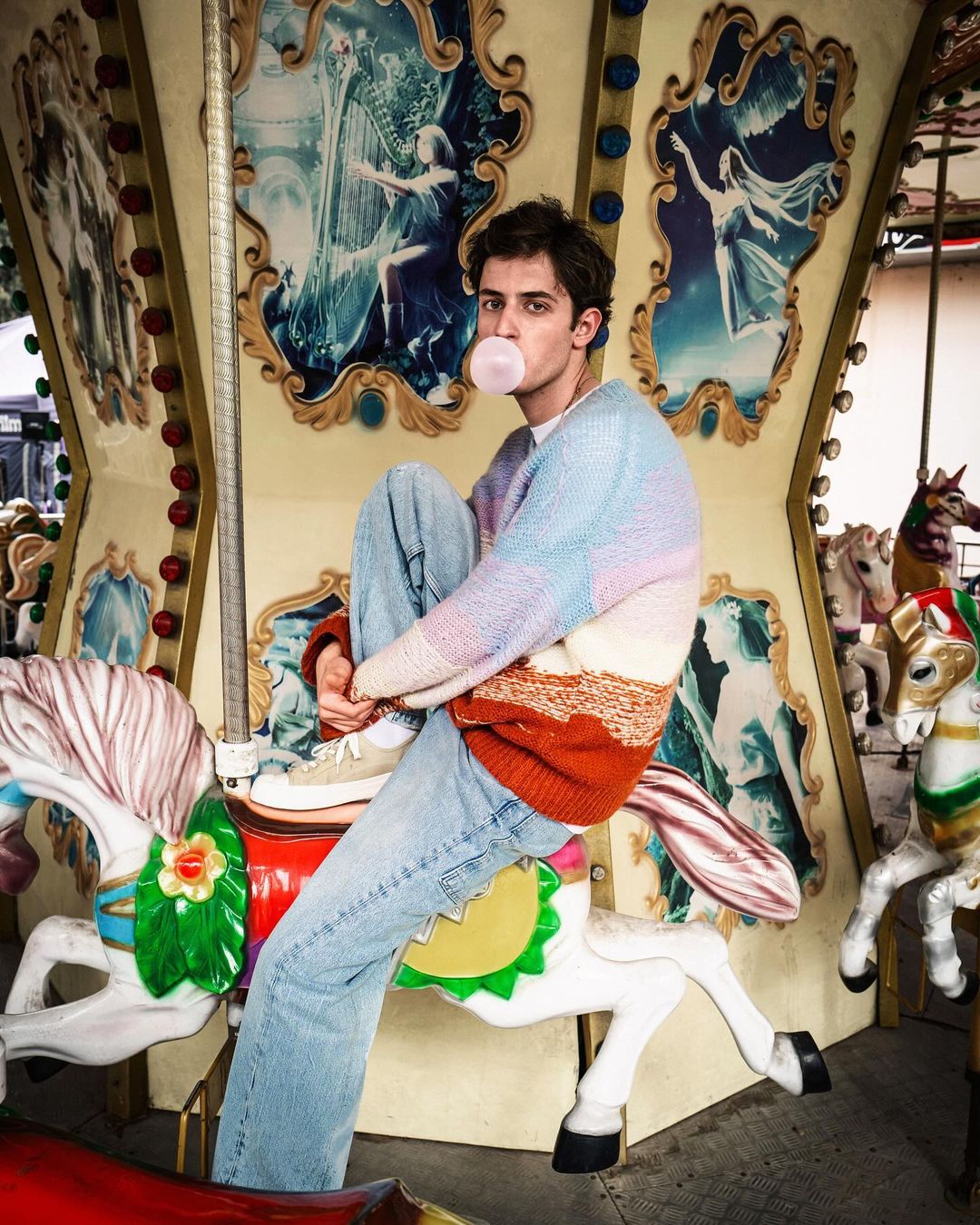 Young adult male blowing bubblegum bubble, seated on carousel horse with light blue jeans and pastel sweater. Candid portrait on fairground.