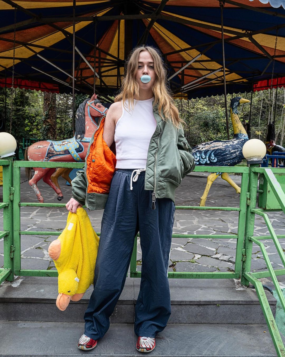 A young woman standing in front of a vintage merry-go-round, blowing a bubble with gum, wearing casual clothing and holding a plush duck.