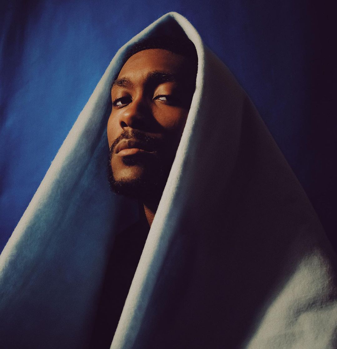 Portrait of a black adult male with a beard, wearing a light blue hooded blanket and looking at the camera against a blue background.