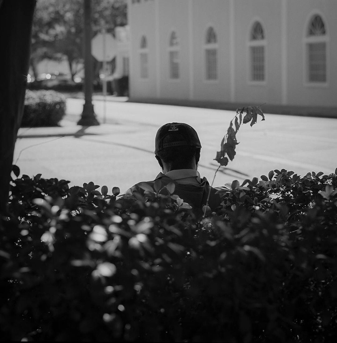 Black and white photo of a man wearing a baseball cap from the back, standing behind green bushes in an outdoor urban setting.