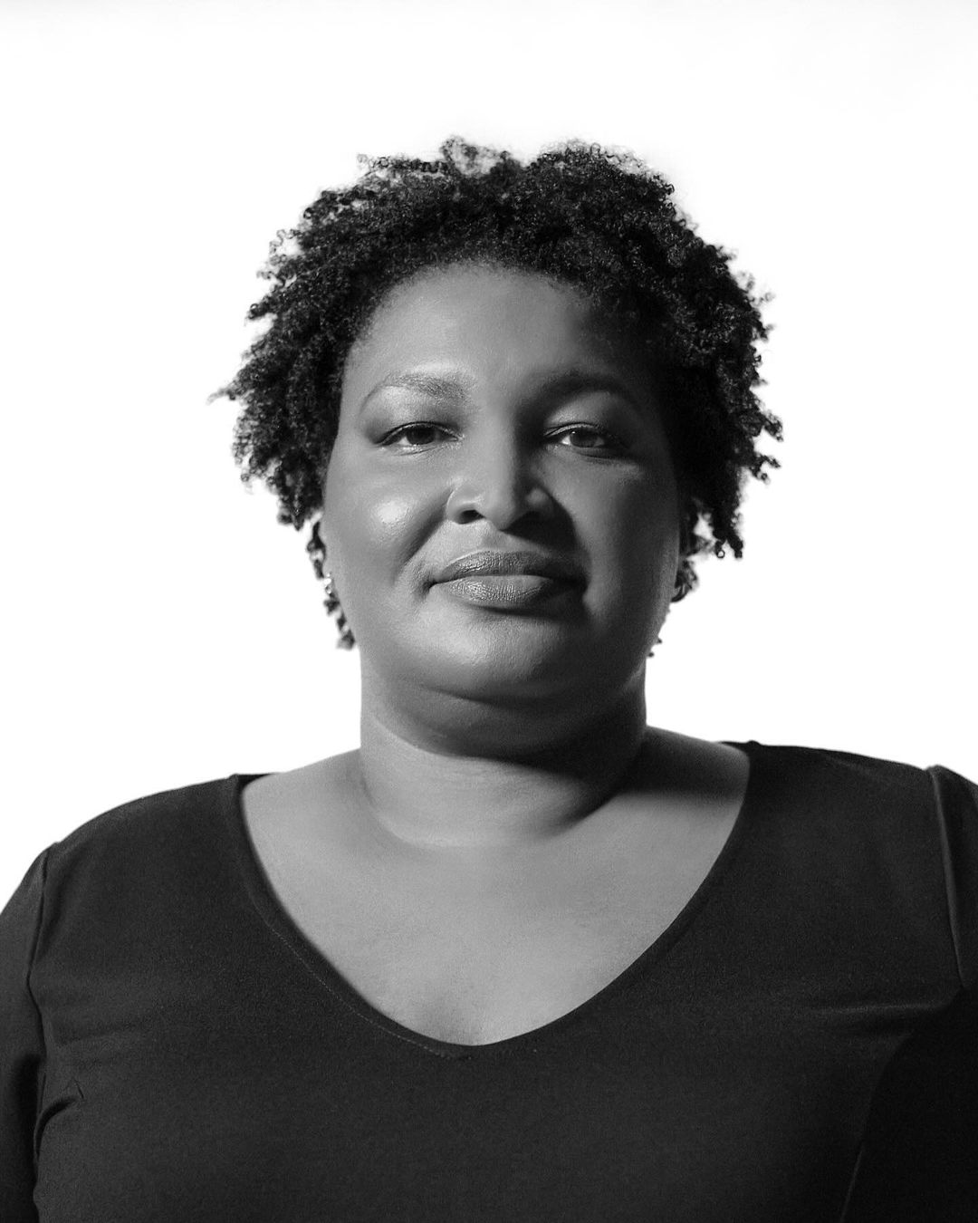 Monochrome portrait of an African-American woman with short natural hair, wearing a dark V-neck top against a bright white backdrop.