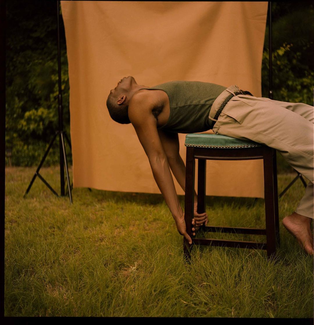 A black man in an olive tank top and khaki pants lies on a green stool outside in grass. A tan backdrop is behind him.