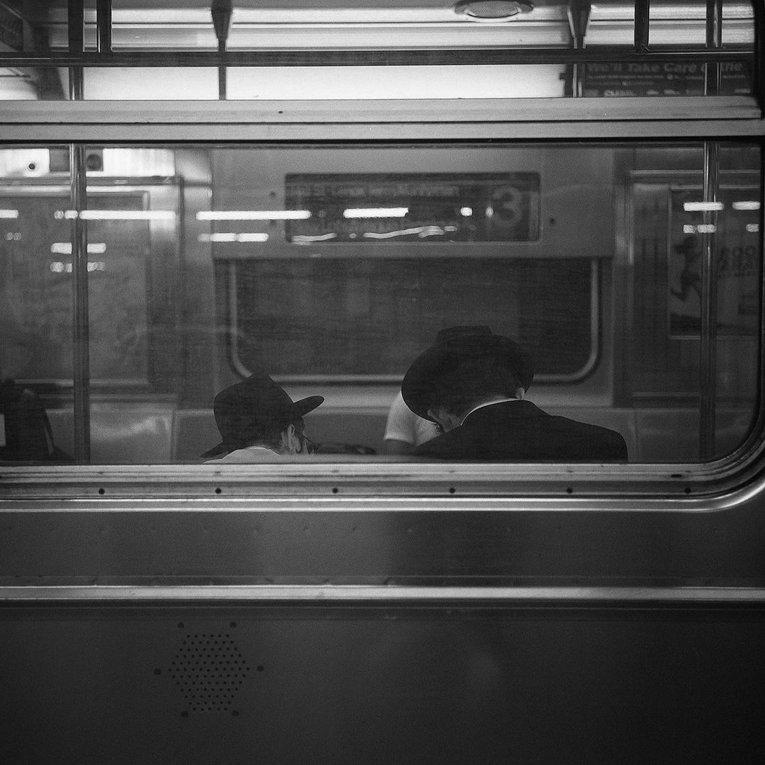 Black-and-white shot inside a subway car featuring two people wearing hats, seen through the train window on a moving public transport.
