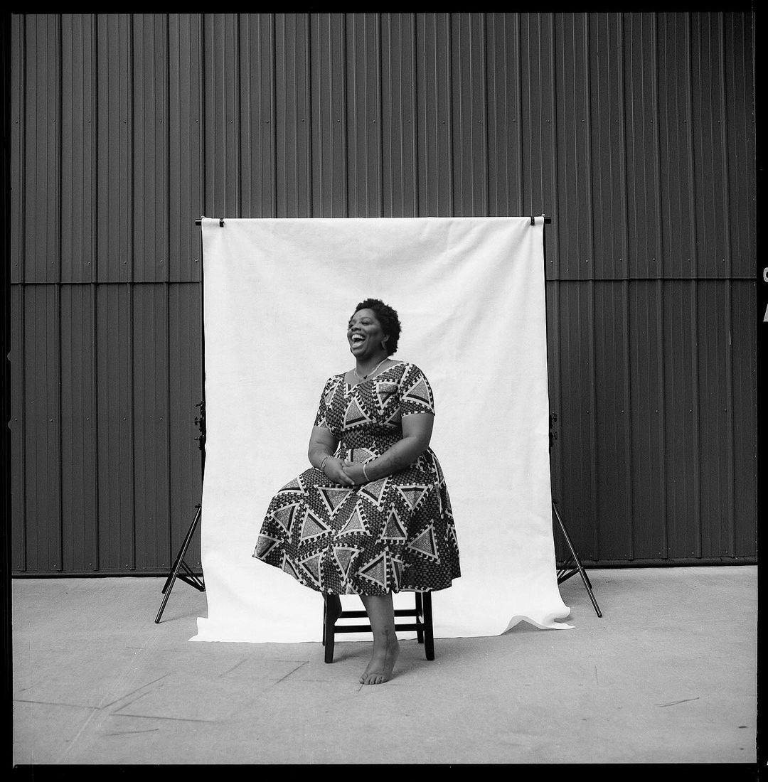 Portrait of a happy adult woman in a patterned dress sitting on a stool in front of a white backdrop outdoors.