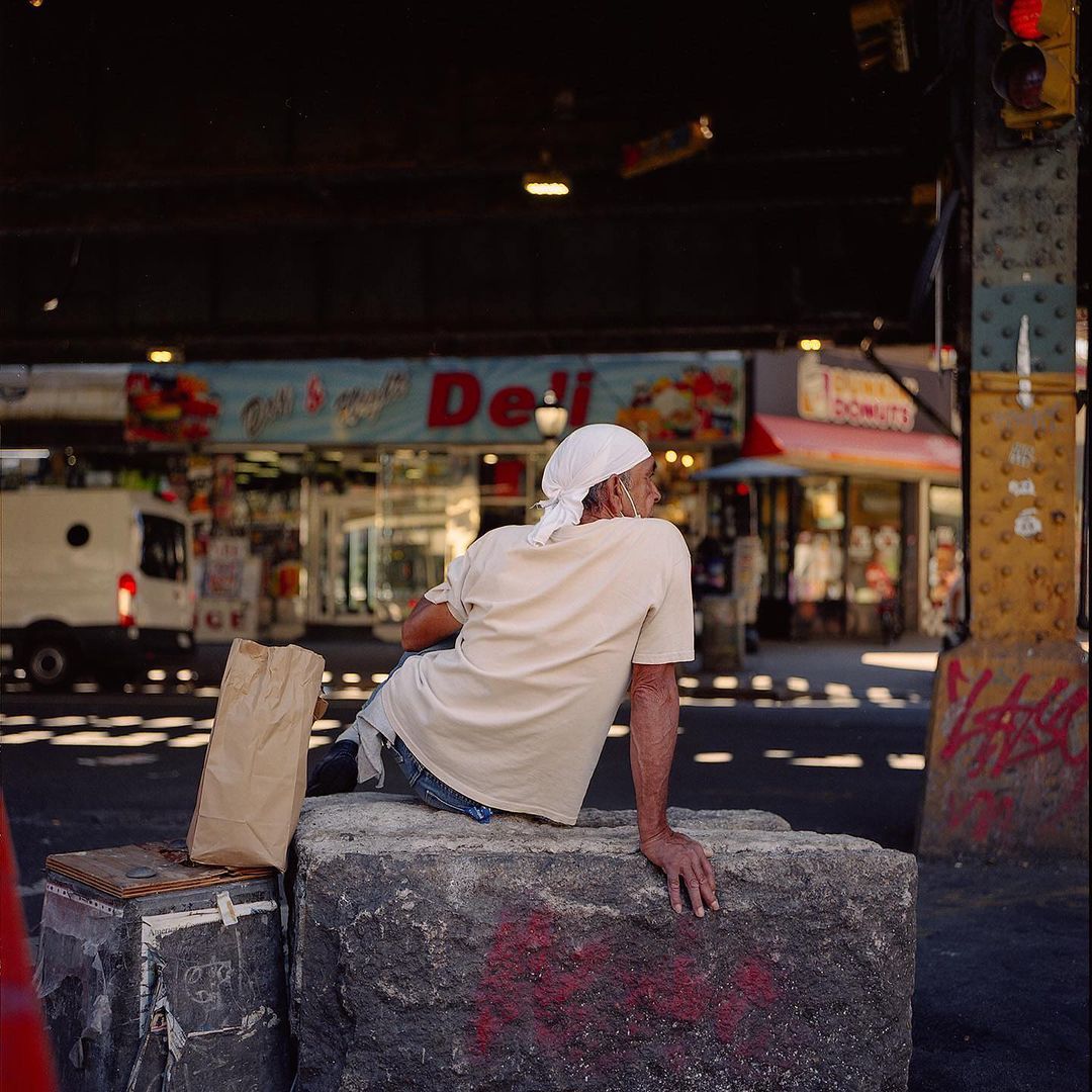 Man resting on concrete blocks under a bridge near a city deli and donut shop on a sunny day, wearing a head covering and casual clothes.