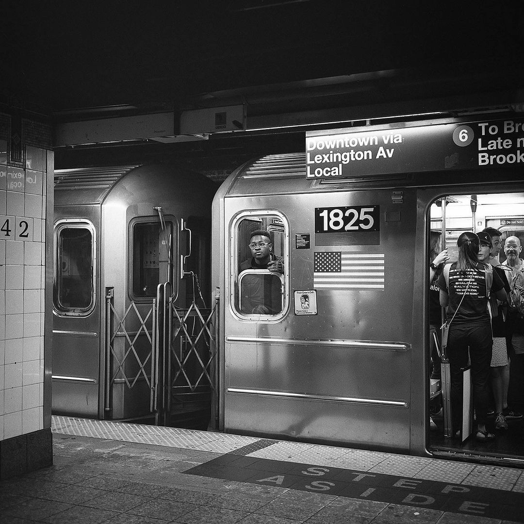 Black and white shot of a subway train car at a station in New York City, with passengers boarding and an MTA sign visible.