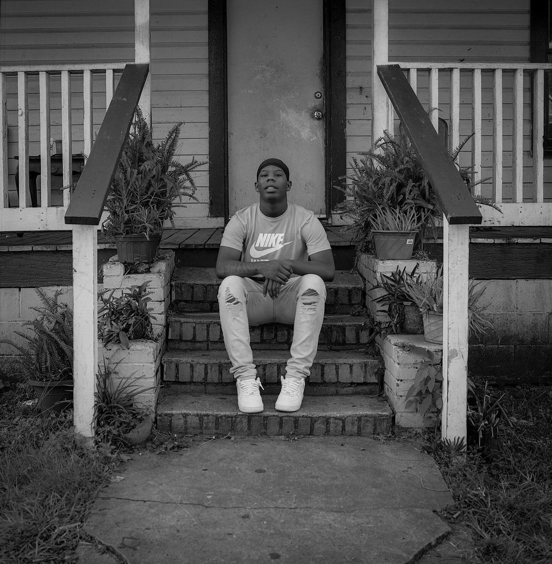 Black and white portrait of a young Black man seated on the steps of a house wearing a t-shirt, ripped jeans, and sneakers.