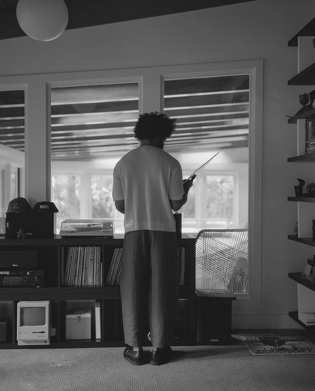 Back view of person with afro holding a device in a room with a shelf, records, and an Apple Macintosh computer.