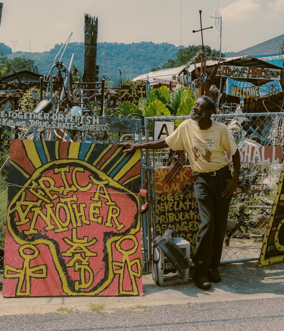 Adult male stands next to an outdoor display of painted signs and artwork. Buildings and trees are visible in the background. 