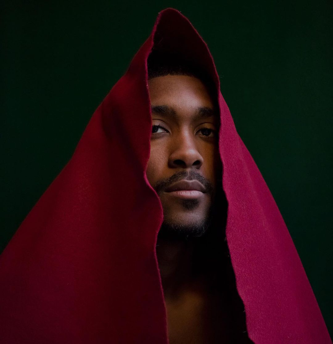 Headshot of a dark-skinned man with a red hood, looking seriously at the camera against a dark green background in a portrait.