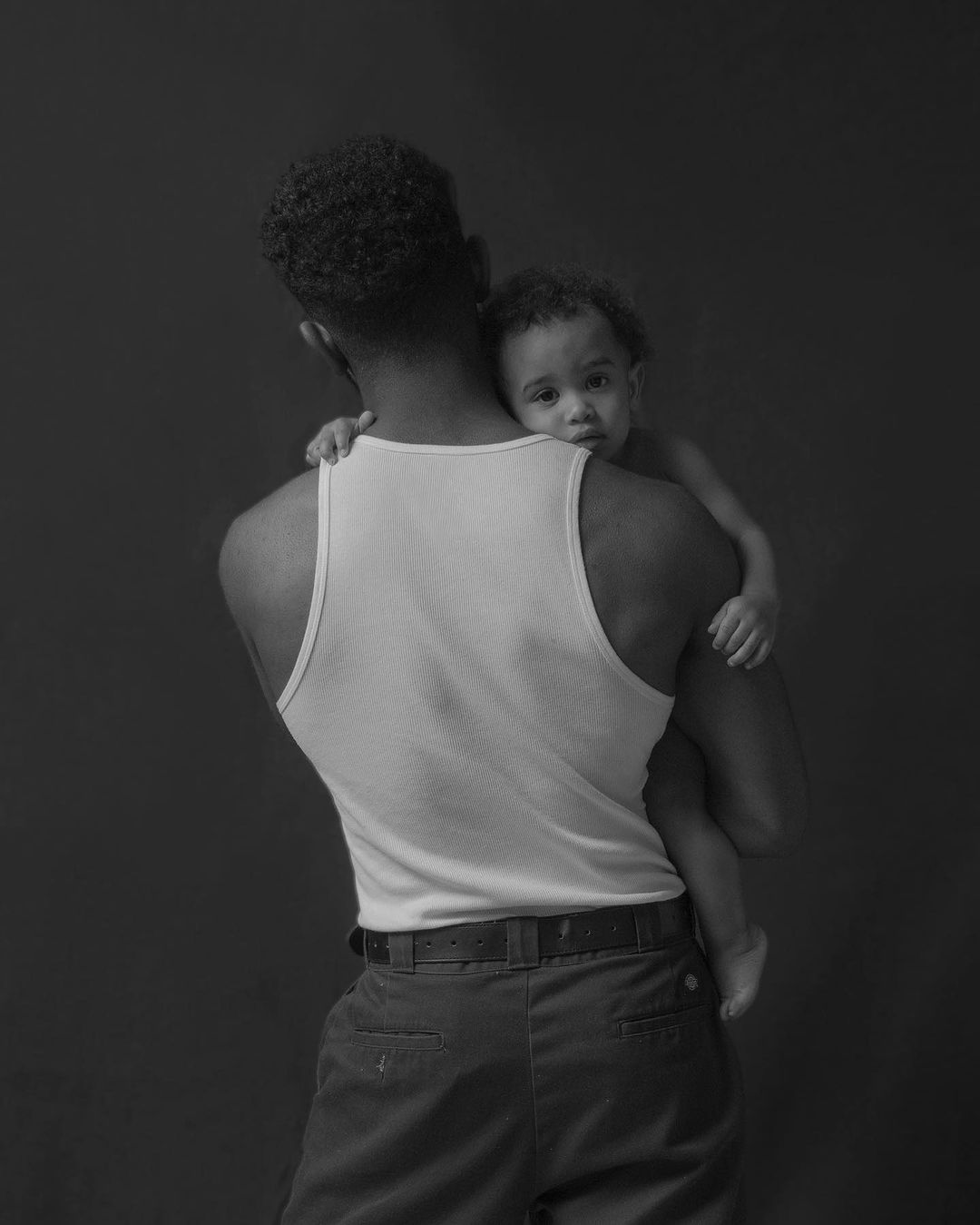 Monochrome portrait of an African-American man holding a baby, both facing away from the camera against a dark backdrop. Fatherhood.