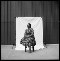 Black woman wearing a patterned dress sitting on a stool in front of a white seamless backdrop outdoors
