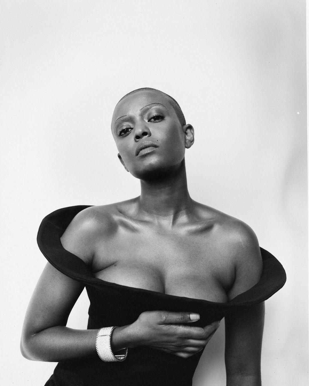 Black and white portrait of a woman with a shaved head wearing a wide-collar dress and a bracelet in a studio setting.
