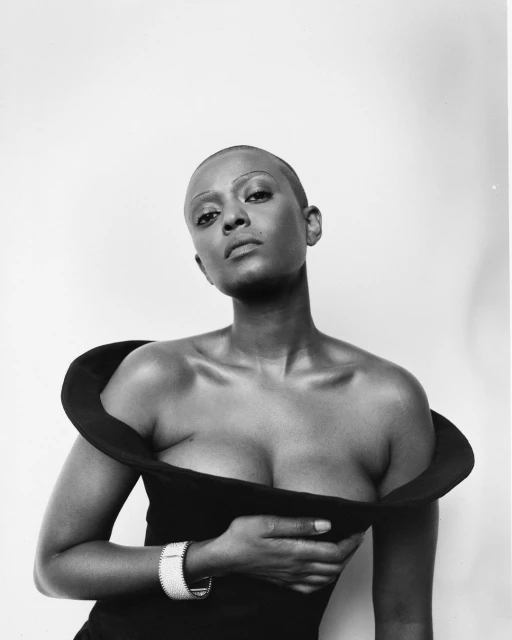 Black and white portrait of a woman with a shaved head wearing a wide-collar dress and a bracelet in a studio setting.