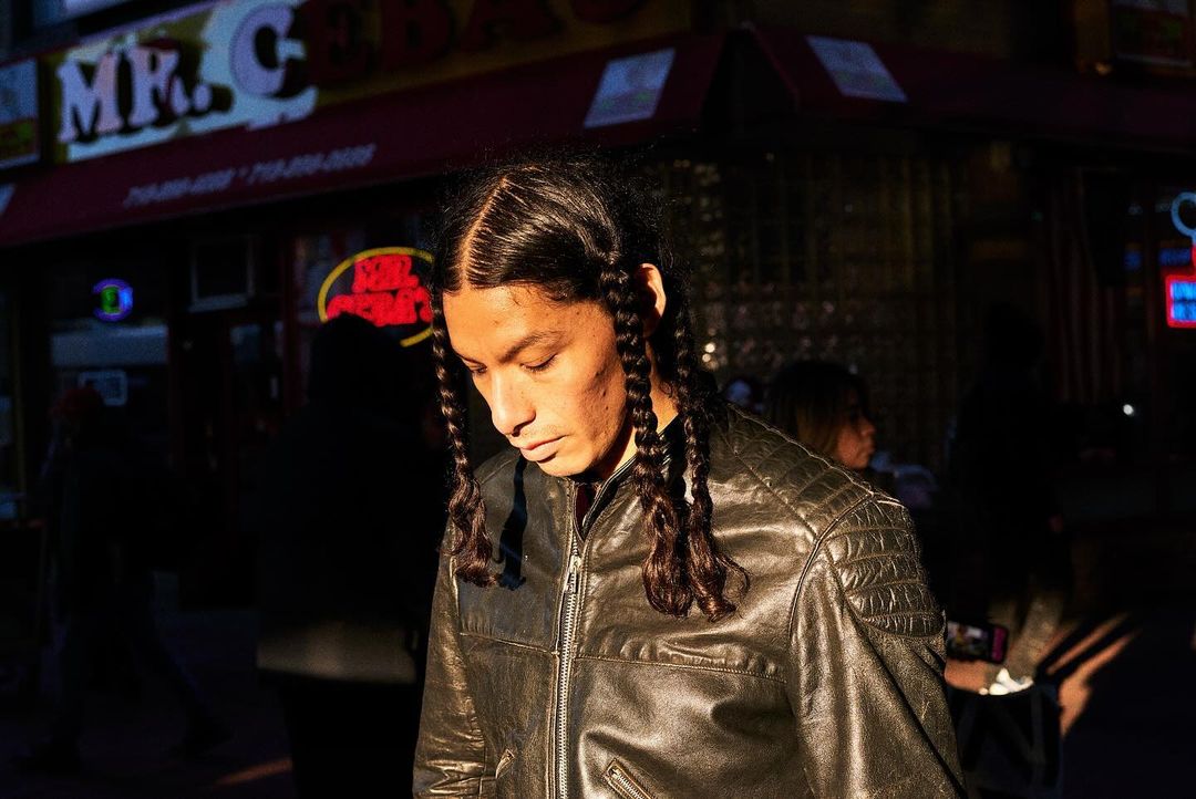 Adult male with braided hair poses in a dark leather jacket on a city street at night with brightly lit store fronts.
