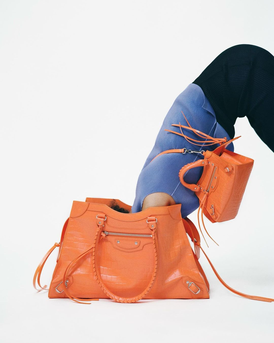 Fashion editorial shot showcasing two textured orange leather Balenciaga purses against a stark white backdrop, featuring a human figure.