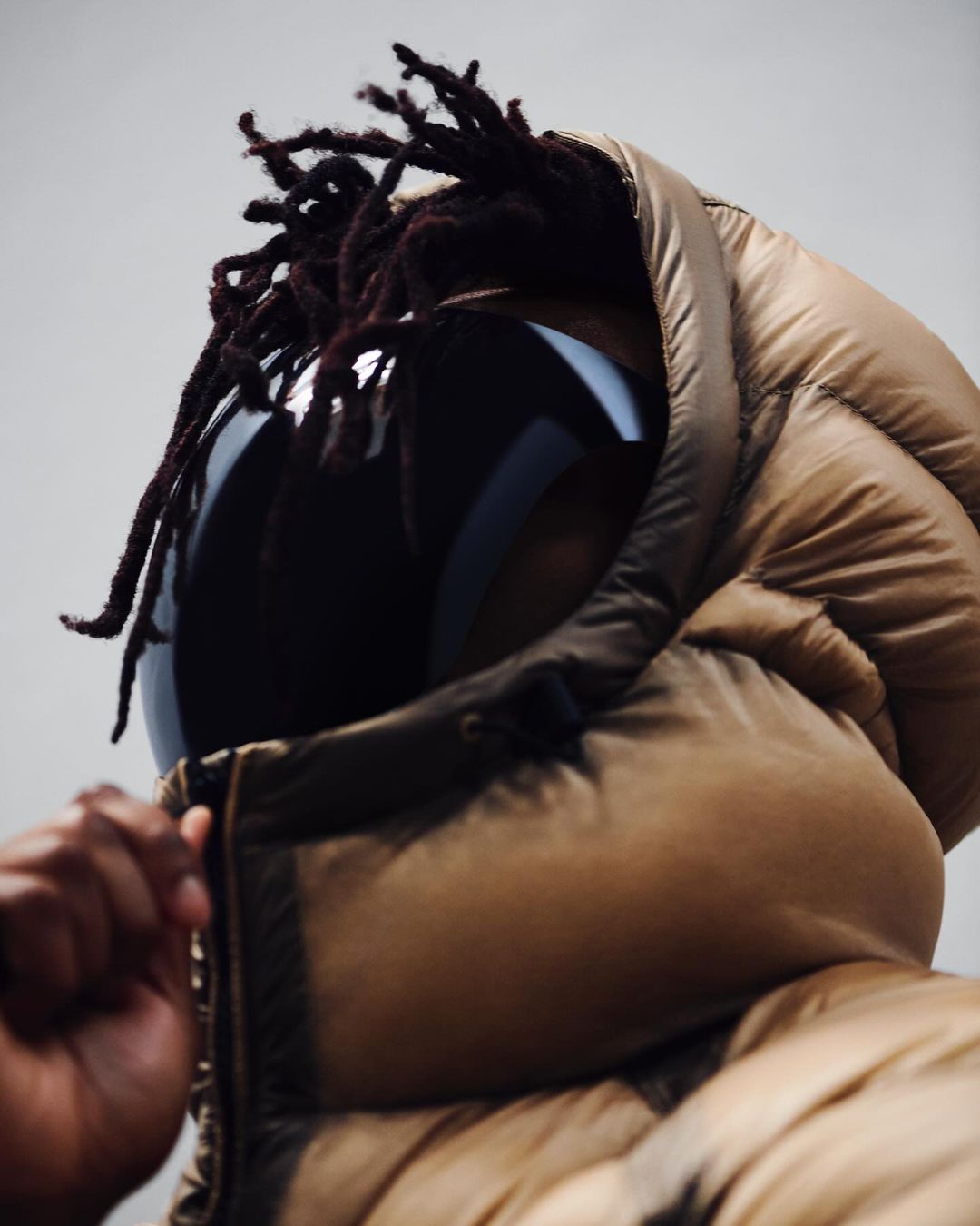 Person in a puffer jacket with the hood up and reflective sunglasses, facing slightly downward against a plain grey background.