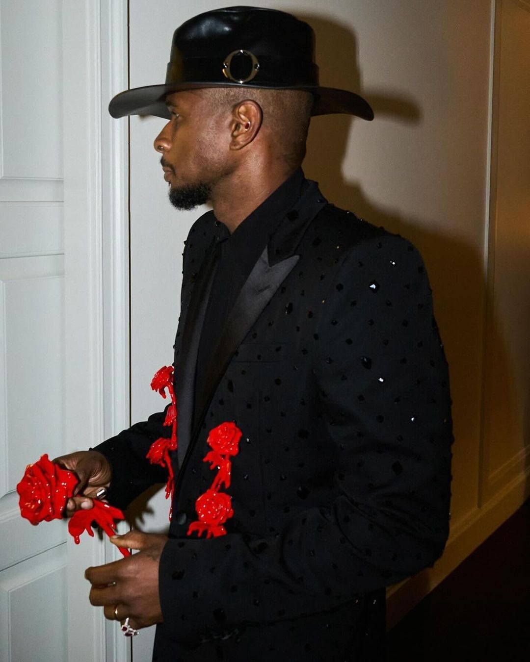 Side profile of a Black male in a black bejeweled suit and wide-brimmed hat, accented with red rose adornments. Set against a neutral indoor background.