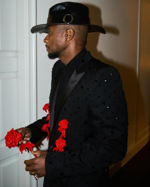 Side profile of a Black male in a black bejeweled suit and wide-brimmed hat, accented with red rose adornments. Set against a neutral indoor background.