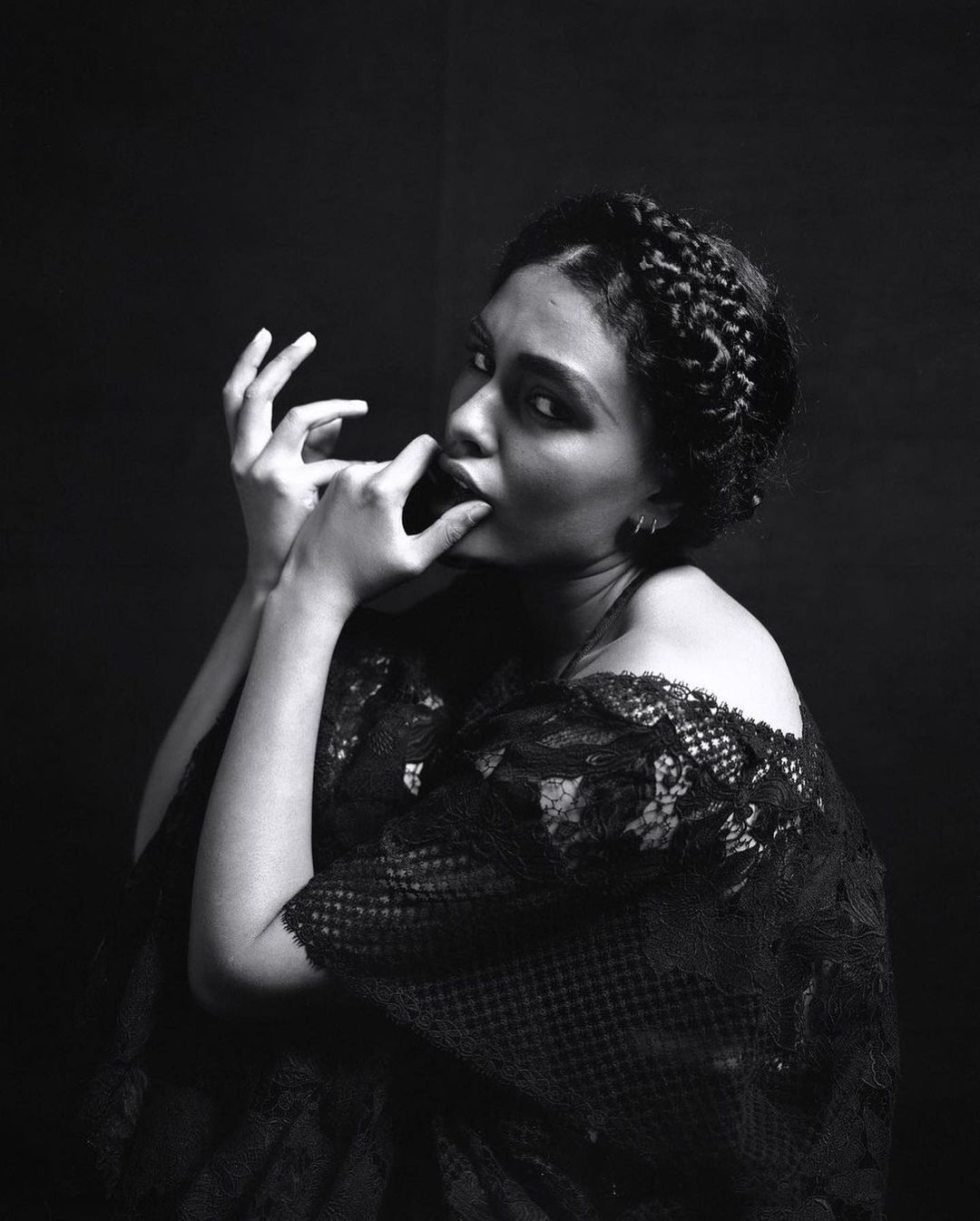 Monochrome portrait of an adult female with dark hair in braids, wearing a black lace top, fingers near her mouth, against a black background.