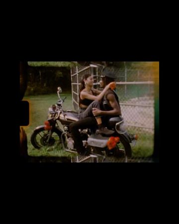 Two people sitting on a parked motorcycle outdoors near a chain-link fence captured in film frame style with sprocket holes.