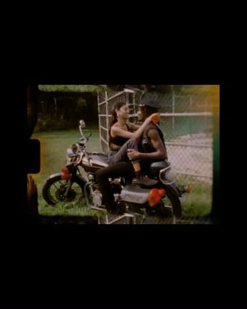 Two people sitting on a parked motorcycle outdoors near a chain-link fence captured in film frame style with sprocket holes.