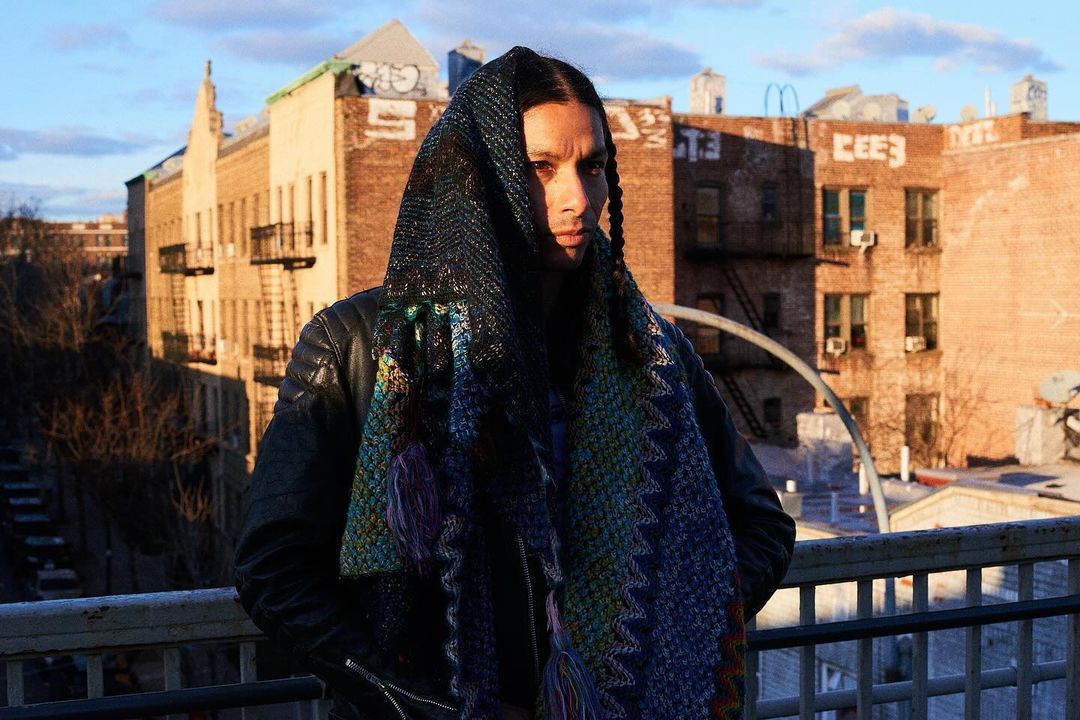 Man wearing patterned hooded scarf and leather jacket on a rooftop with city buildings and blue sky at sunset.