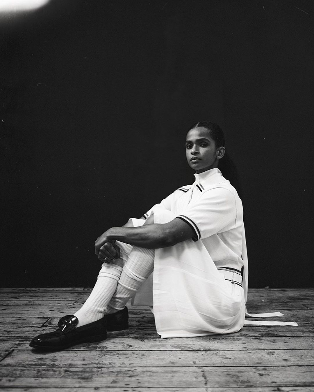 Monochrome portrait of a person seated indoors wearing a white tunic, white socks, and black leather shoes against a dark backdrop.