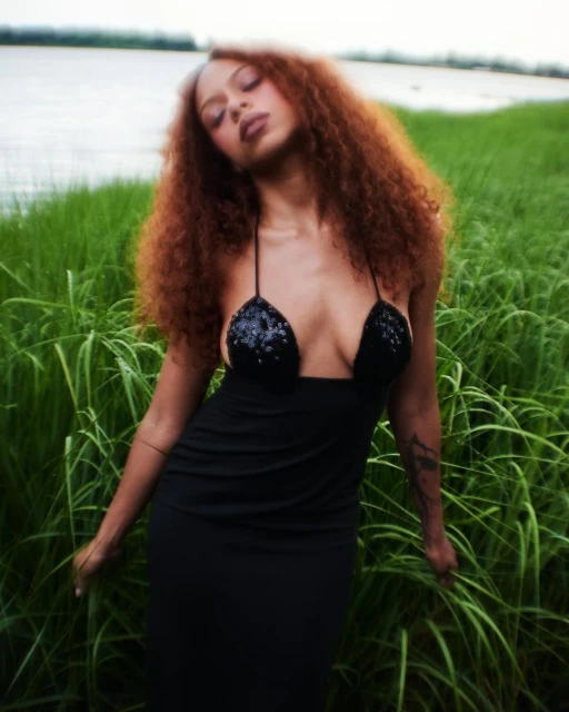 A woman with auburn curly hair, wearing a black dress, stands in a field of tall green grass with a blurry body of water in the background.