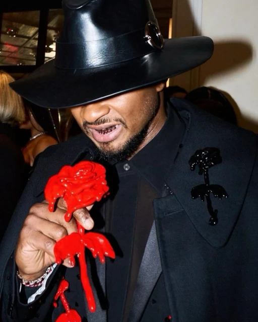 Usher poses indoors wearing a black hat, cape, and shirt while holding a red rose; his smile reveals gold teeth grills.