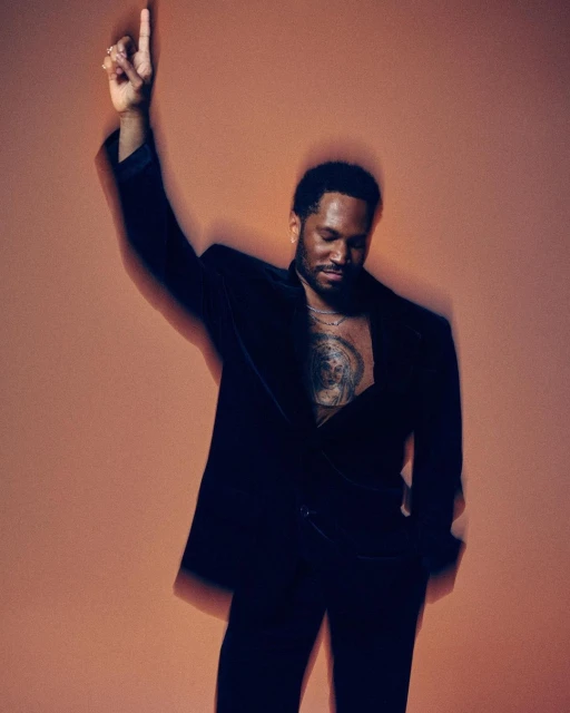 Portrait of Black male model in open velvet blazer pointing upward against an orange backdrop in a brightly lit photo studio.
