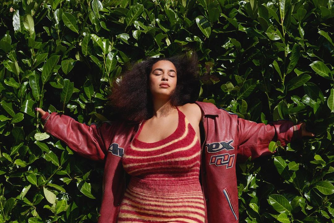 Adult woman with dark curly hair in red striped dress and jacket posing outdoors against green foliage background with eyes closed.