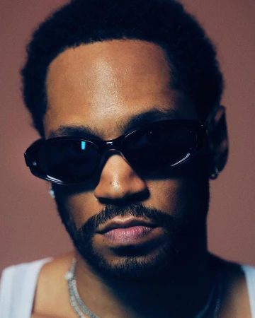 Close-up portrait of a Black man wearing dark sunglasses, a white tank top, and layered silver chain necklaces.