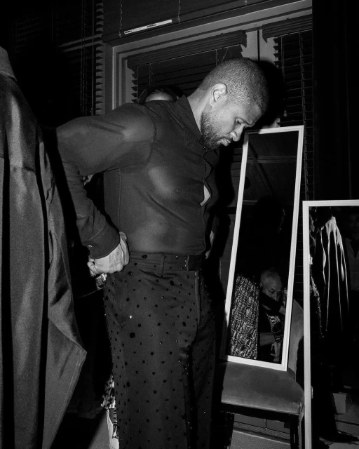 Man adjusts pants in a monochrome image with mirrors reflecting other people and clothes rack on the side creating an intimate backstage scene.