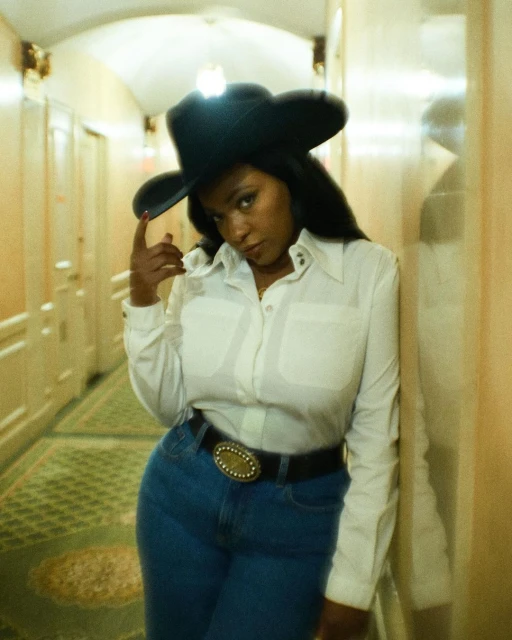 Portrait of a woman in a cowboy hat, white shirt, and jeans in a hotel hallway, leaning against the wall with soft, diffused lighting.
