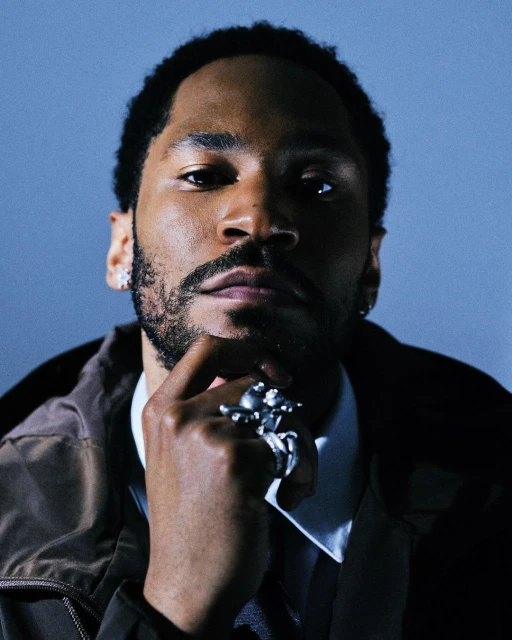 Portrait of a Black man in a jacket and collared shirt, wearing a statement ring, with a cool blue background. Head and shoulders.