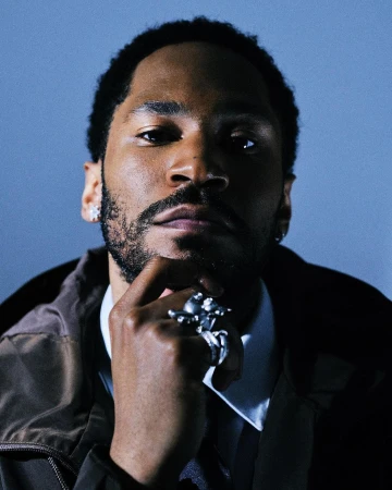 Portrait of a Black man in a jacket and collared shirt, wearing a statement ring, with a cool blue background. Head and shoulders.