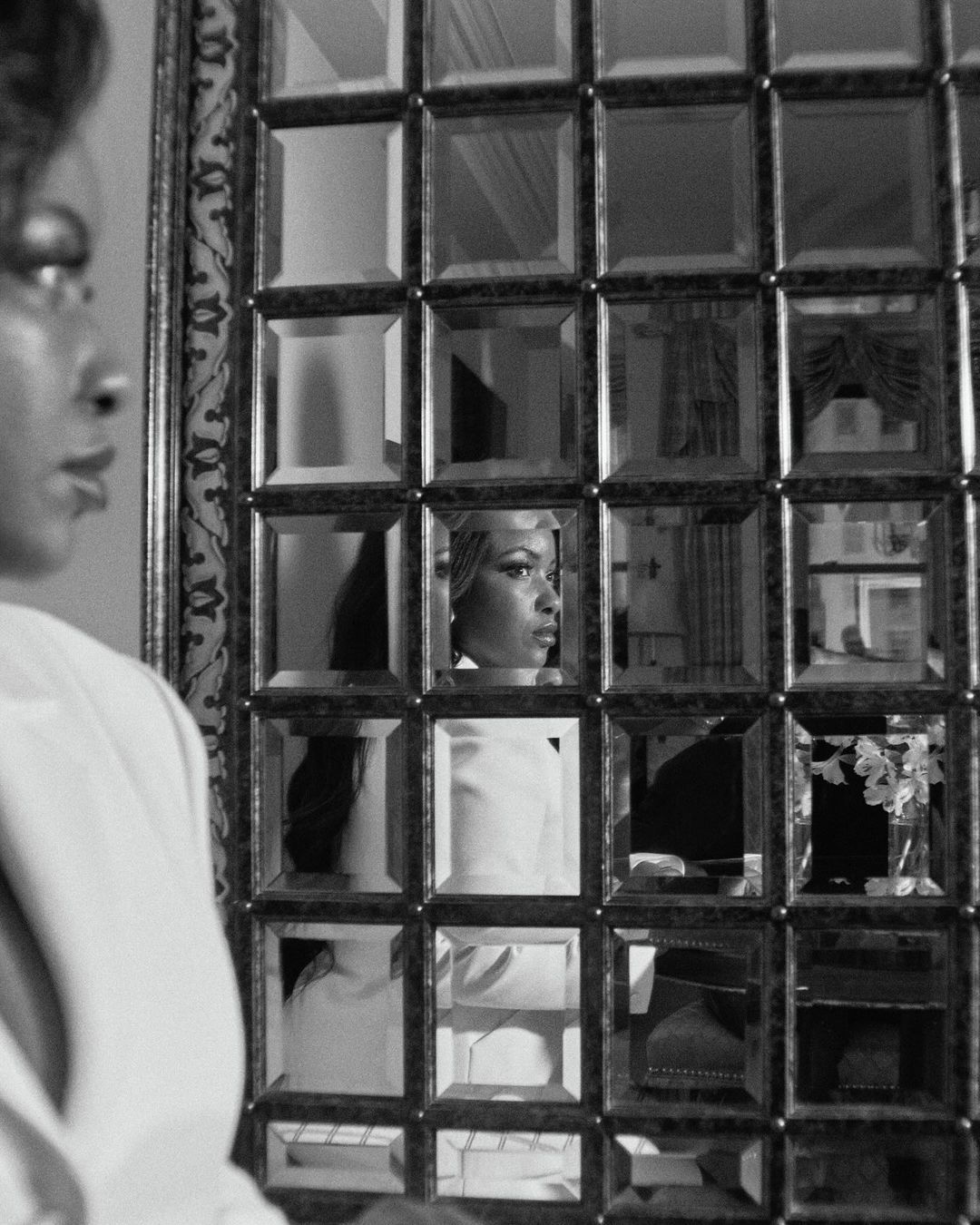 Monochrome portrait of an African-American woman in white formalwear, partially reflected in a grid-patterned decorative mirror.