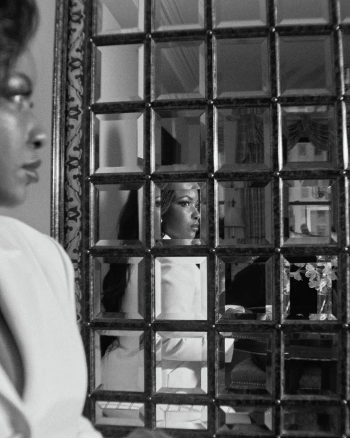 Monochrome portrait of an African-American woman in white formalwear, partially reflected in a grid-patterned decorative mirror.